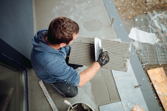 Portrait professionnel du dirigeant ou de l'équipe dans le showroom de l'entreprise, entouré d'échantillons de carrelage, faïence et revêtements de sol exposés. L'atmosphère doit être chaleureuse et professionnelle, montrant l'accueil client, avec des éléments du showroom visibles en arrière-plan (présentoirs de carrelage, calepins de devis, matériaux d'aménagement). La personne doit être souriante et accessible, incarnant la disponibilité et l'expertise d'une entreprise familiale établie depuis longtemps.