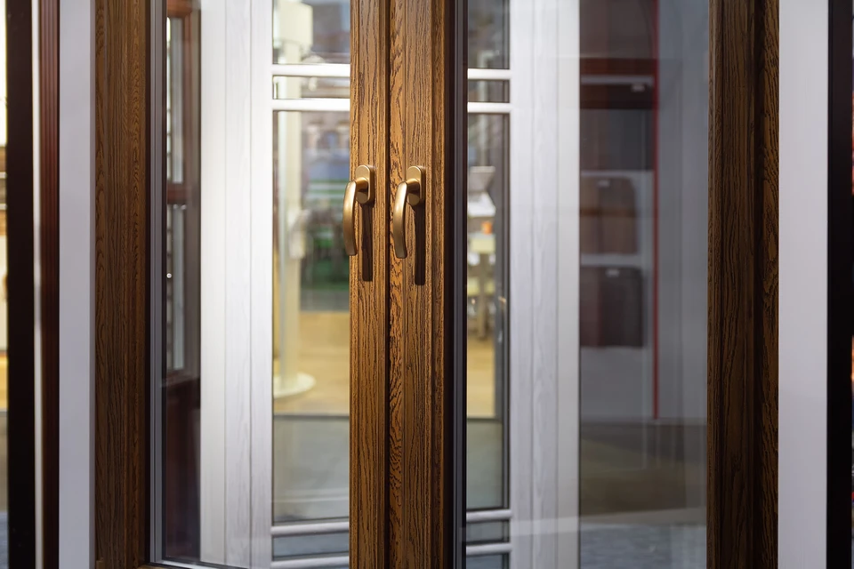 Showroom de menuiseries et revêtements intérieur : espace lumineux et accueillant exposant des fenêtres et portes de différents styles, coloris et matériaux (PVC, aluminium, bois) disposées ou installées sur des cloisons de présentation. Au premier plan, détail de portes d'entrée modernes avec poignées visibles. Ambiance professionnelle et chaleureuse avec éclairage mettant en valeur les produits, permettant au client de visualiser concrètement les finitions et teintes disponibles.